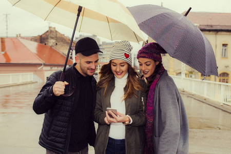 三个快乐而微笑的朋友用手机拿着雨伞