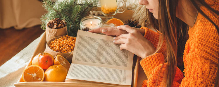 柔软舒适的照片, 妇女在温暖的橙色毛衣在床上与一杯茶和水果。女孩坐在床上与旧书