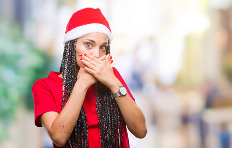 年轻的编织头发非洲裔美国女孩戴着圣诞帽在孤立的背景震惊覆盖嘴与手的错误。秘密概念