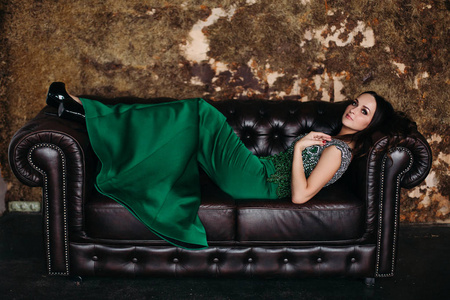 华丽美丽的女人在绿色的礼服躺在黑色的皮沙发上