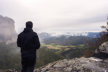 有遮挡的人从一个秋天的山谷的悬崖上注视着地平线上阴云密布的山脉, 山谷被雾覆盖着, 在背景中你可以看到农田