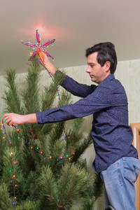 英俊的中年男子装饰一棵圣诞树与一颗星和一个五颜六色的灯泡的花环站在楼梯上的客厅内。温馨家居新年假期理念