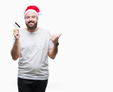 年轻的高加索人戴着圣诞帽拿着信用卡在孤立的背景指向和显示与幸福的脸微笑的边