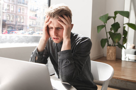 惊呆了的年轻人坐在一家小咖啡馆里, 看着笔记本电脑, 双手举起来照片