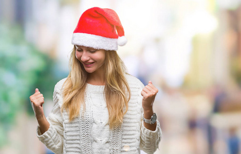 年轻美丽的金发女子戴着圣诞节帽子在孤立的背景非常高兴和兴奋做赢家手势与手臂举起, 微笑和尖叫的成功。庆典理念