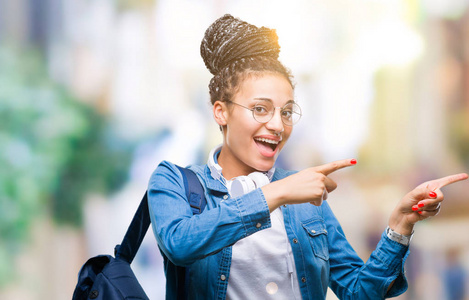 年轻的编织头发非洲裔美国学生女孩在孤立的背景上背着背包微笑着, 看着相机指向镜头, 用两只手和手指指着一边