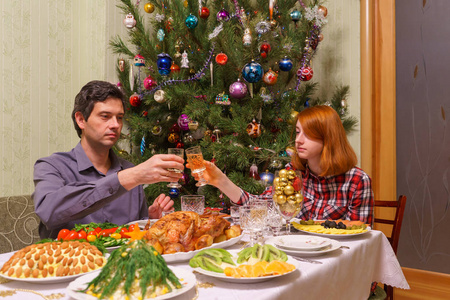 英俊的中年男子和美丽的年轻女士在客厅内部装饰圣诞树附近的节日餐桌上喝着香槟酒庆祝新年假期。舒适的家居理念