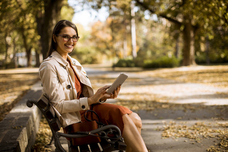 妇女坐在公园长凳在秋季天气使用平板电脑和检查社交媒体