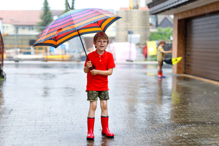 小男孩穿着红色的雨鞋, 在城市街道上带着五颜六色的雨伞散步。孩子在夏天的时候戴着眼镜。愉快的孩子在繁忙的夏天阵雨雨