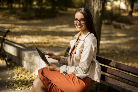 妇女坐在公园长凳在秋季天气使用平板电脑和检查社交媒体