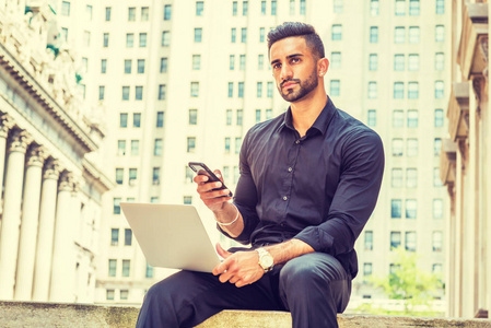 成功之路。年轻的东印度裔美国人, 留胡子的人在纽约街头工作, 穿着黑色衬衫, 拿着笔记本电脑, 坐在旧式办公楼外, 用手机发短信