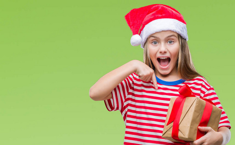 年轻美丽的女孩戴着圣诞帽子和持有礼物在孤立的背景与惊讶的脸指向自己的手指