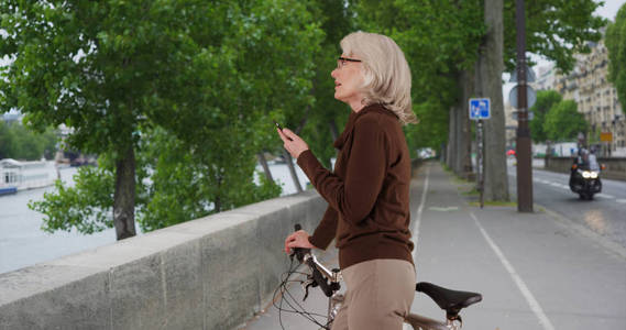 活跃的高级白种人妇女骑自行车看着河流和电话发短信