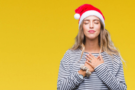 美丽的年轻金发女子戴着圣诞节帽子在孤立的背景微笑与手在胸部与闭的眼睛和感恩的手势在脸上。健康理念