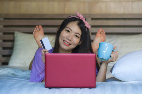 年轻美丽和快乐的亚洲日本妇女使用信用卡网上银行在笔记本电脑上的笔记本电脑在家里微笑着愉快的购物网上购买电子商务和生活方式的概念