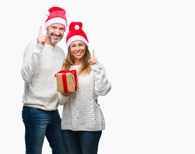中年拉美裔夫妇穿着圣诞帽子和持有礼物在孤立的背景惊讶的想法或问题指向手指与幸福的脸, 第一