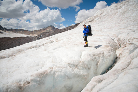 戴着帽子和太阳镜的旅行者, 肩膀上的背包放在冰川上的雪山上, 对着天空和云层。自然环境中的旅行者