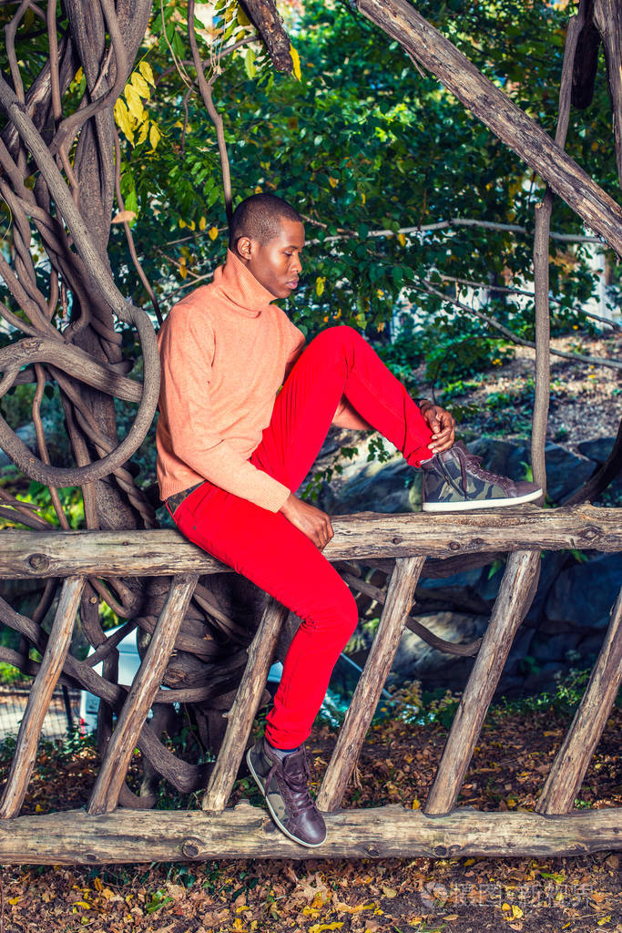 红色裤子, 图案靴子鞋, 一个年轻的黑人坐在栅栏上, 弯曲的腿, 向下