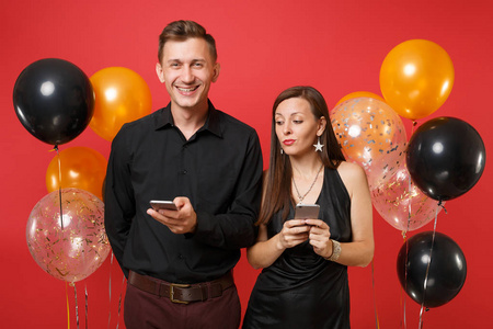 夫妇在黑色的衣服短信在手机庆祝生日假日派对孤立在红色背景气球。情人节国际妇女节新年快乐2019概念。模拟