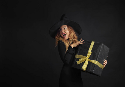 黑色星期五。万圣节女巫与黑色的帽子在黑色背景拿着礼物盒与朱斯在她的手中。惊喜和礼品在售