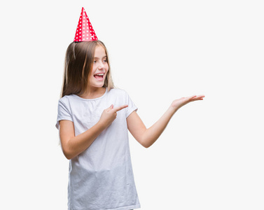 年轻美丽的女孩戴着生日帽在孤立的背景惊讶和微笑的镜头, 同时提出用手和手指指着
