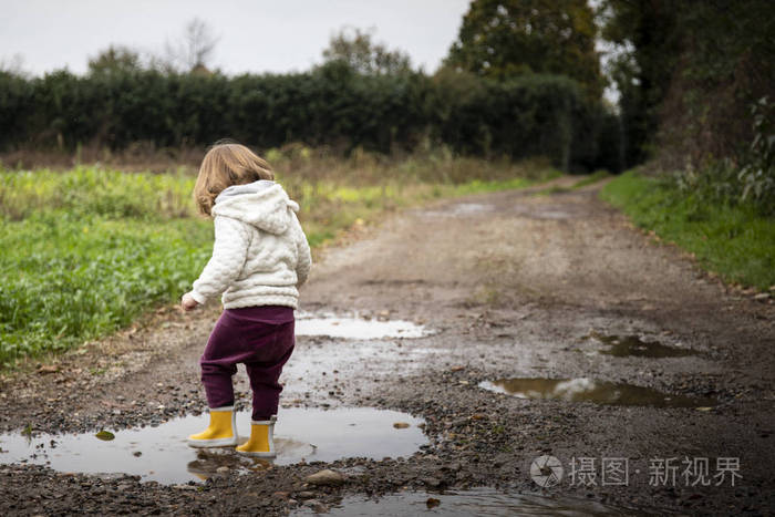 幼儿女孩, 后景, 在泥泞的乡村路上, 用黄色雨靴在水坑里飞溅.