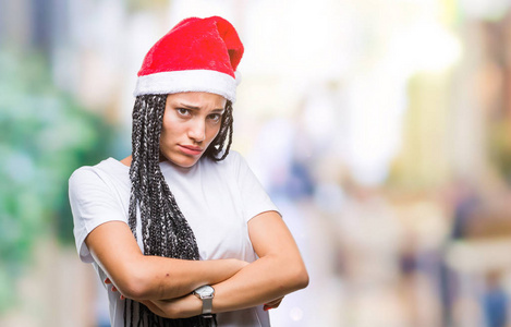 年轻的编织头发非洲裔美国女孩戴着圣诞帽在孤立的背景怀疑和紧张, 不赞成的表情在脸上交叉的武器。消极的人