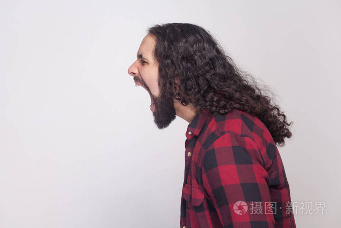 侧面视图英俊愤怒的男人与胡子和黑色长卷发在格子红色衬衫站立和尖叫