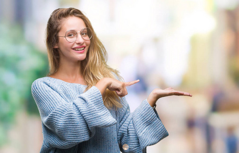 年轻美丽的金发女子穿着毛衣和眼镜在孤立的背景惊讶和微笑的相机, 而用手和手指指着