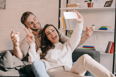 幸福的年轻夫妇采取自拍与智能手机, 并在家里的沙发上作出和平姿态