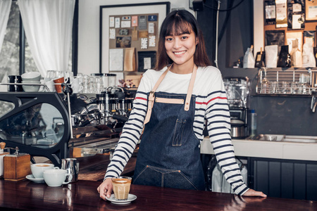 亚洲女店员穿吉恩围裙将她的手置于柜台栏和微笑给客户，咖啡馆的服务理念，所有者商业启动