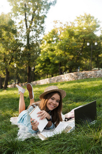 迷人的年轻少女躺在公园的草地上, 学习与笔记本电脑和书籍