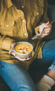 女性穿着黄色衬衫和牛仔裤坐在和吃秋天温暖黄色南瓜奶油汤与面包从杯与勺子。秋季素食, 素食主义者, 健康舒适食品饮食的概念