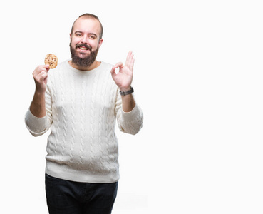年轻的嬉皮士男子吃巧克力片饼干在孤立的背景做确定的标志与手指, 优秀的象征