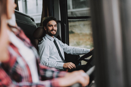 微笑的人驾驶旅游巴士。专业司机。年轻的快乐男子穿着白色衬衫坐在驾驶座上, 看着女人。有吸引力的自信的人在工作。旅游理念