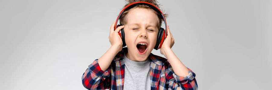 一个英俊的男孩穿着格子衬衣, 灰色衬衫和牛仔裤站在灰色的背景上。一个戴着红色耳机的男孩唱着歌