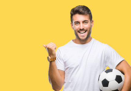 年轻英俊的男子拿着足球足球在孤立的背景指向和显示与拇指到一边, 快乐的脸微笑