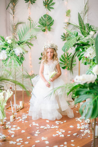 可爱的小女孩, 金发碧眼的卷发在白色的婚纱礼服和花圈的花朵装饰品, 由热带树叶, 白玫瑰和兰花