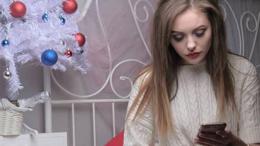 一个穿着白色针织毛衣的美丽的长发金发女孩坐在床上悲伤地看着电话, 同时等待新的一年。圣诞树, 一瓶酒和彩球在背景上
