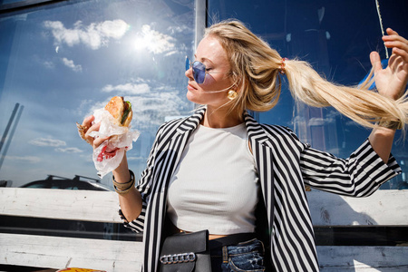 可爱的金发女孩的肖像戴太阳镜, 条纹夹克和白色 t恤吃三明治户外