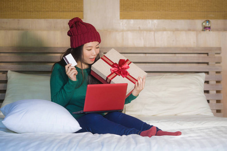 年轻美丽和快乐的亚裔美国妇女在家床上拿着信用卡和圣诞礼物盒购物网上圣诞礼物微笑满意电子商务和电子商务的概念