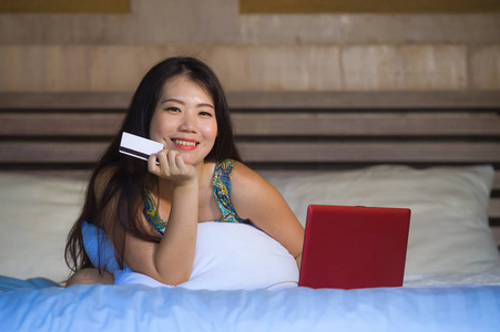 年轻美丽和快乐的亚洲韩国妇女使用信用卡网上银行在笔记本电脑上的家在床上微笑兴奋的购物网上购买电子商务和生活方式的概念