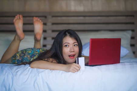 年轻美丽和快乐的亚裔美国女孩使用信用卡网上银行在笔记本电脑上的家在床上微笑兴奋的购物网上购买电子商务和生活方式的概念