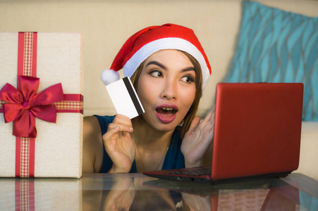 年轻的快乐和美丽的女孩放松在家里的沙发上圣诞老人帽子使用笔记本电脑支付圣诞礼物与信用卡微笑在网上购物和互联网商务