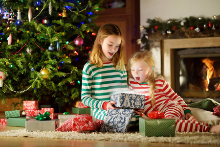 圣诞前夜, 穿着圣诞睡衣的快乐小妹妹们在舒适黑暗的起居室里, 在壁炉旁打开礼品盒。在家庆祝圣诞节