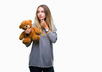 年轻美丽的金发女子抱着泰迪熊毛绒在孤立背景严重的脸思考问题, 非常混淆的想法