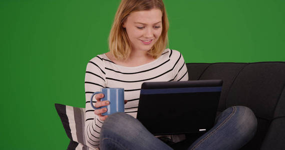 金发碧眼的女孩与咖啡坐在沙发上浏览网页上的笔记本电脑上的绿色屏幕