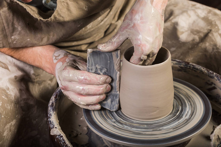 从扭曲的陶工车轮上的粘土创建杯。在粘土和波特砂轮与工件的脏手。在车间雕刻家使水罐出陶器特写