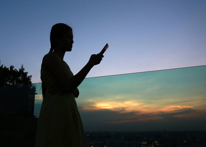 女人在日落时分在建筑物的屋顶上使用智能手机的剪影
