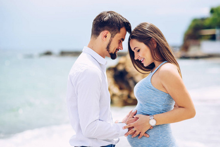 恩爱夫妻上海滩触摸爱与关怀的岩石和海背景上的孕妇的肚子在阳光灿烂